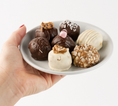 A hand holding a white serving plate with an assortment of 7 Loada Balls protein balls on it with a white background.